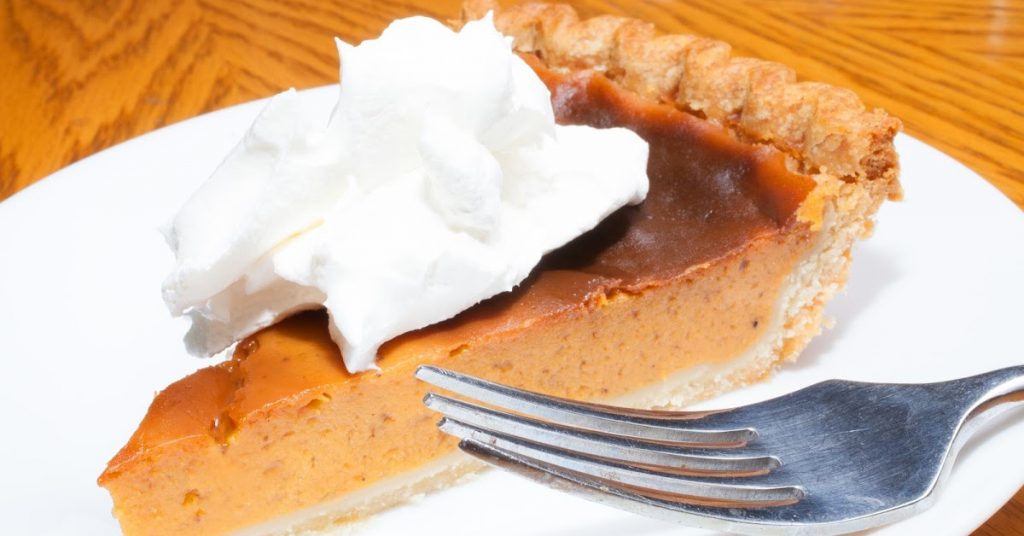 A slice of pumpkin pie topped with a dollop of whipped cream sits on a white plate, with a silver fork resting beside it. The pie has a golden-brown crust and rich, creamy filling. The wooden table underneath adds a warm, rustic touch to the scene.