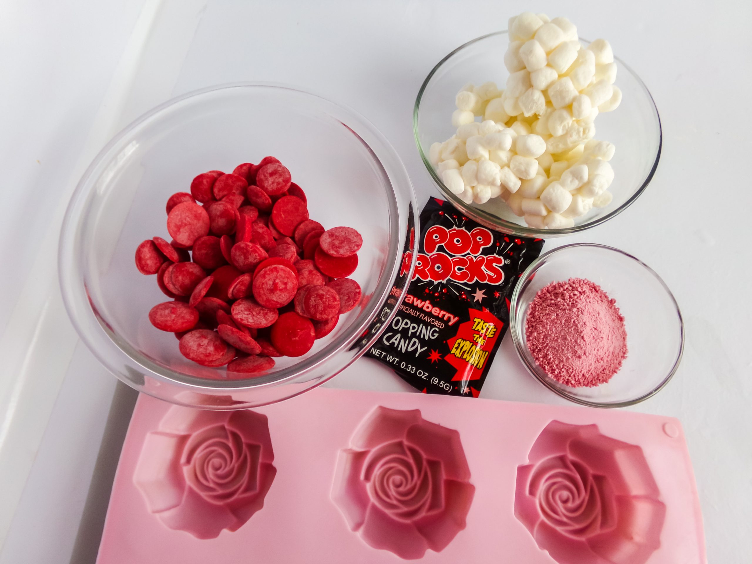 a bowl of red candy melts, a bowl of mini marshmallows, a packet of strawberry Pop Rocks (with the contents poured into a small bowl), and a rose-shaped silicone mold