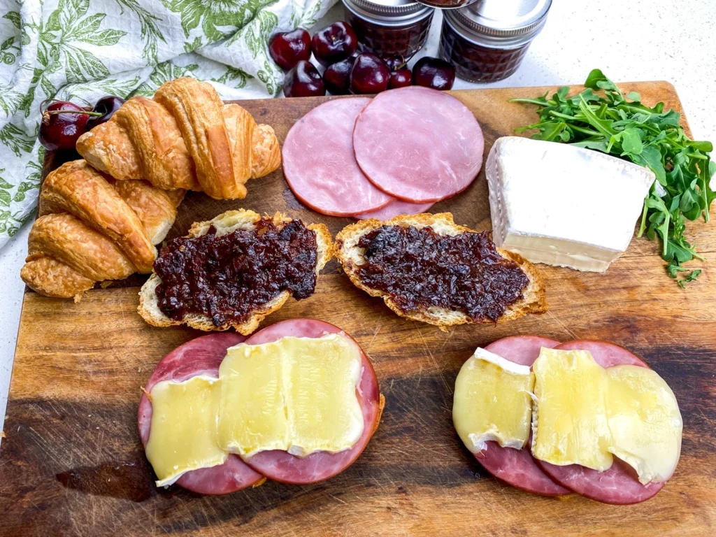 Warm open-faced ham and melted Brie sandwiches with cherry chutney served on a wooden board with fresh ingredients.
