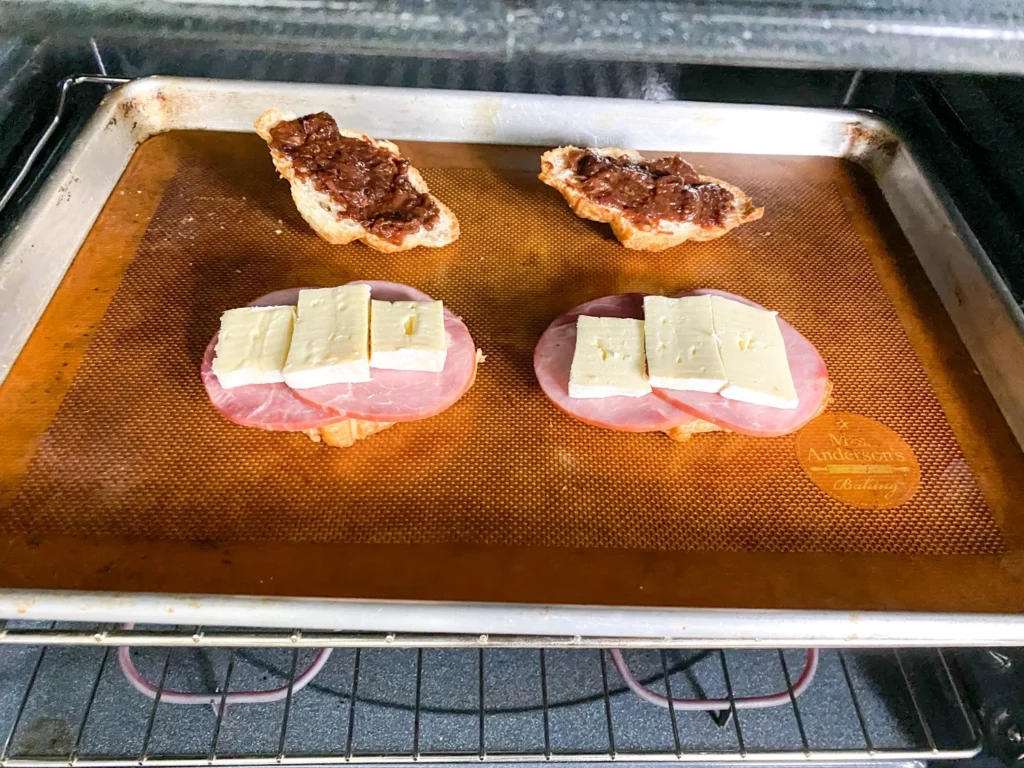 Open-faced ham, Brie, and cherry chutney sandwiches baking on a sheet pan until the cheese melts.