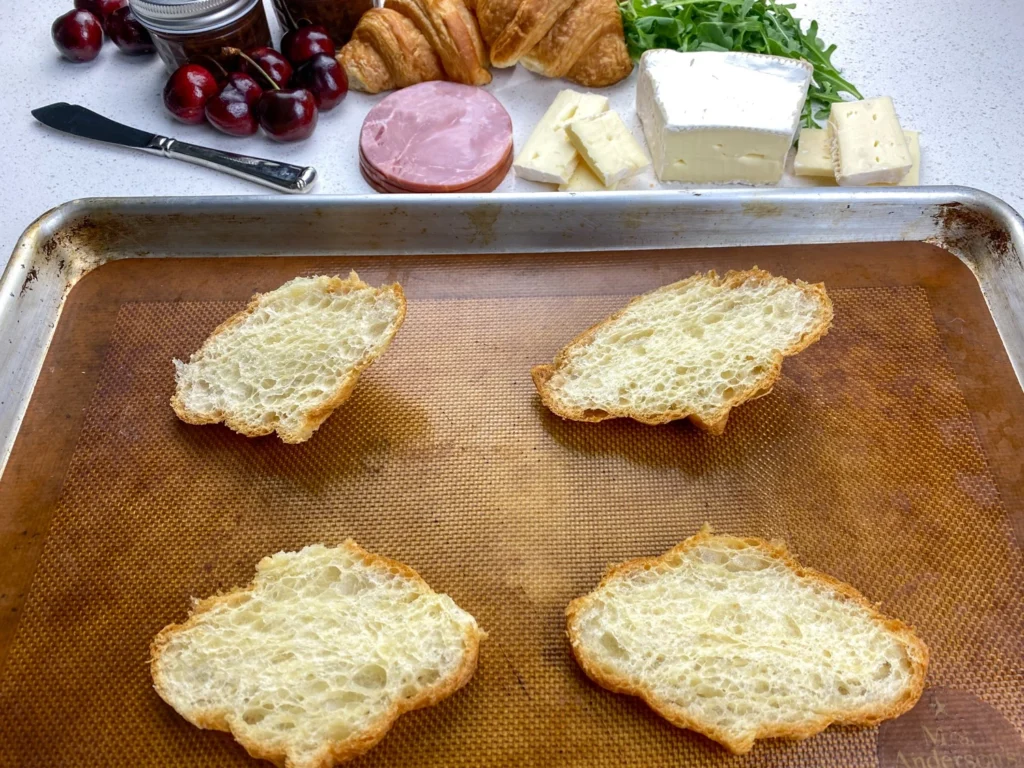 Four slices of artisan bread arranged on a baking sheet to begin assembling ham, Brie, and cherry chutney croissant sandwiches.