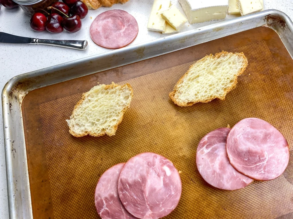 Slices of Black Forest ham placed on toasted artisan bread during assembly of ham and Brie croissant sandwiches.