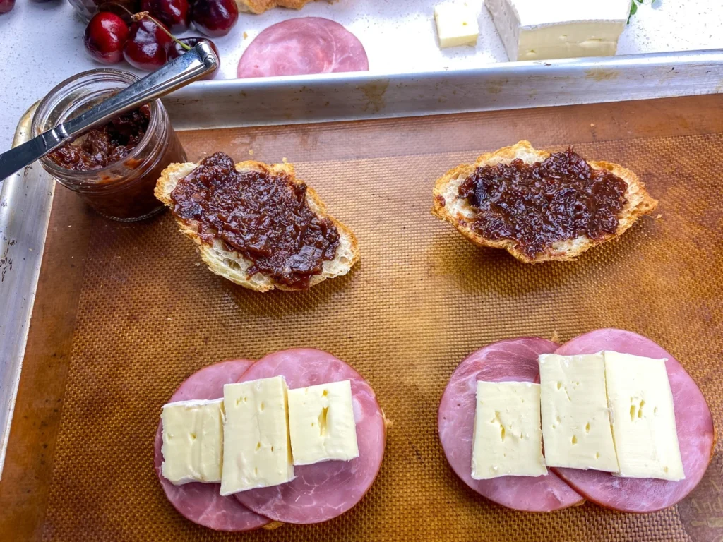Cherry chutney spread onto toasted bread beside open-faced ham and Brie sandwiches ready for the oven.