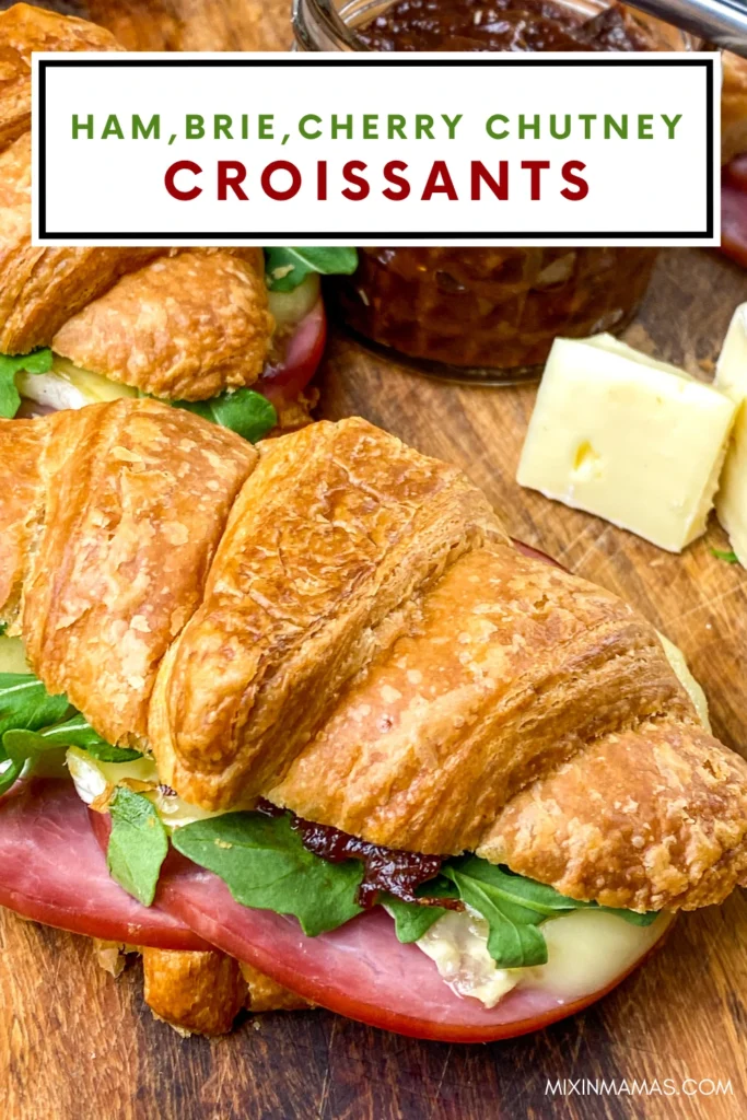 Close-up Pinterest graphic of a ham, Brie, cherry chutney, and arugula croissant sandwich on a wooden serving board.