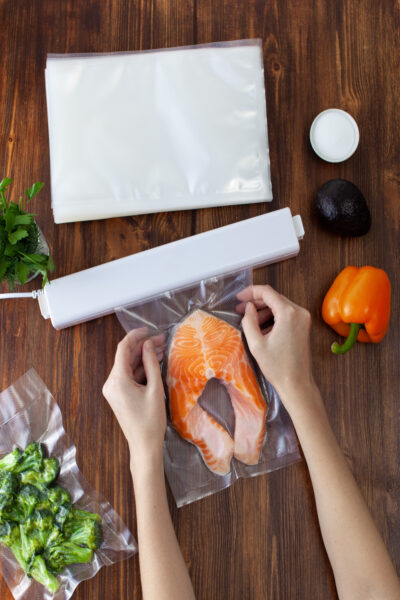 Someone holds a plastic bag with fresh fish inside up to a vacuum sealer device. More bags and ingredients lie around the device.