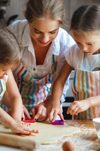 A mom and her two daughters wear matching aprons as they use cookie cutters on dough to cut out cookies.
