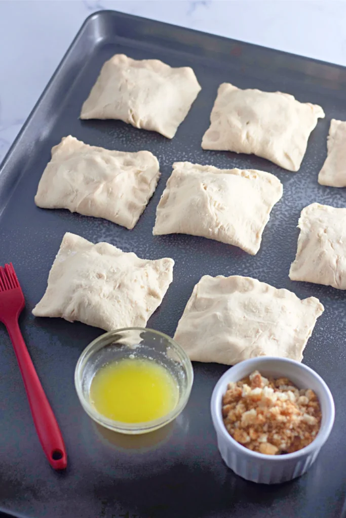 Unbaked chicken crescent pockets on a tray with butter and crushed croutons.