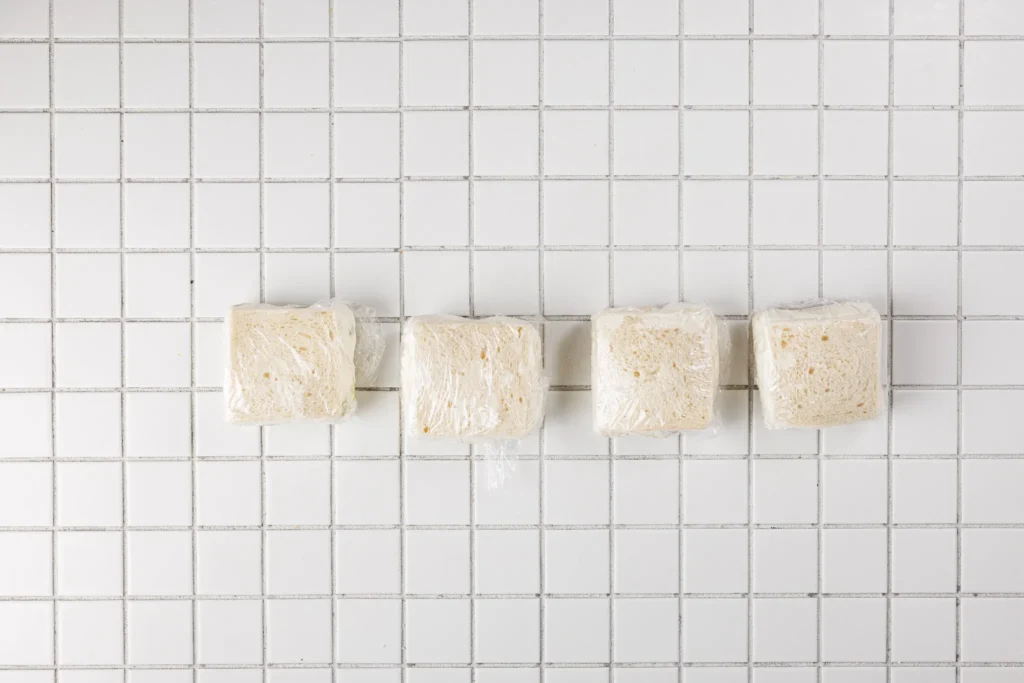 Four Japanese fruit sandwiches wrapped in plastic wrap and lined up on a tiled surface.