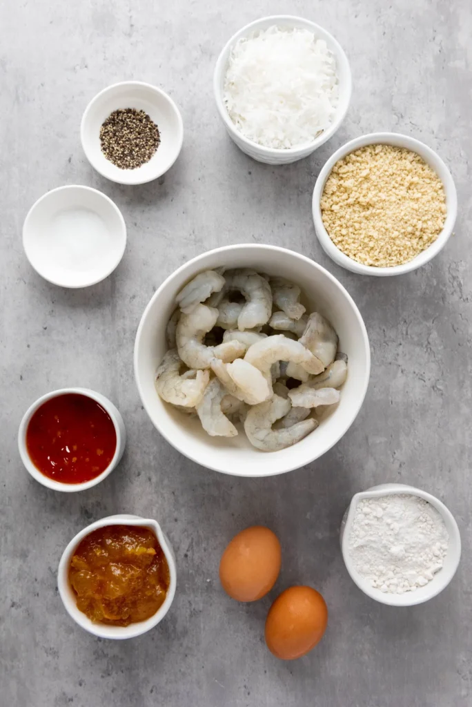 Bowls of shrimp, shredded coconut, panko breadcrumbs, flour, eggs, spices, orange marmalade, and chili sauce.