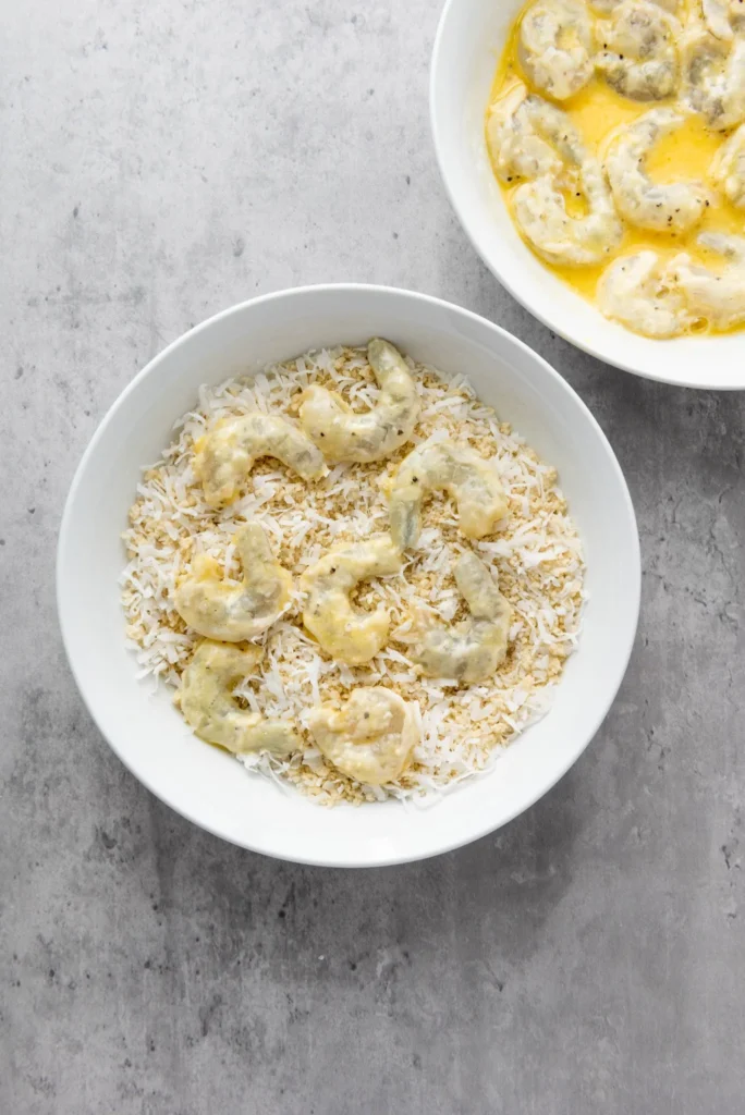 Raw shrimp being coated in shredded coconut and panko breadcrumbs.