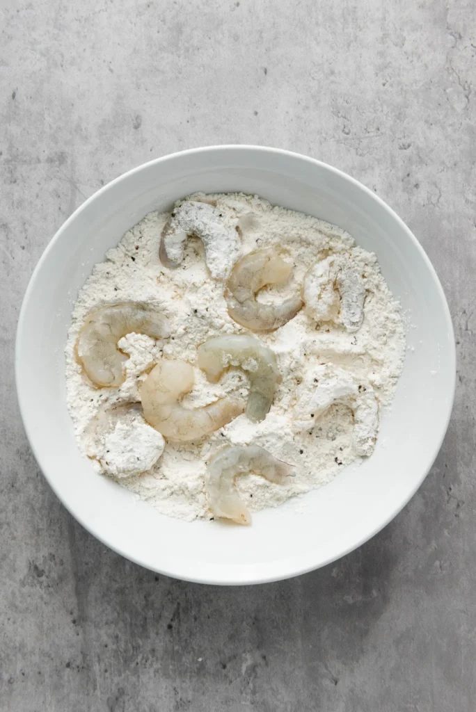 Raw shrimp dredged in seasoned flour in a shallow bowl.