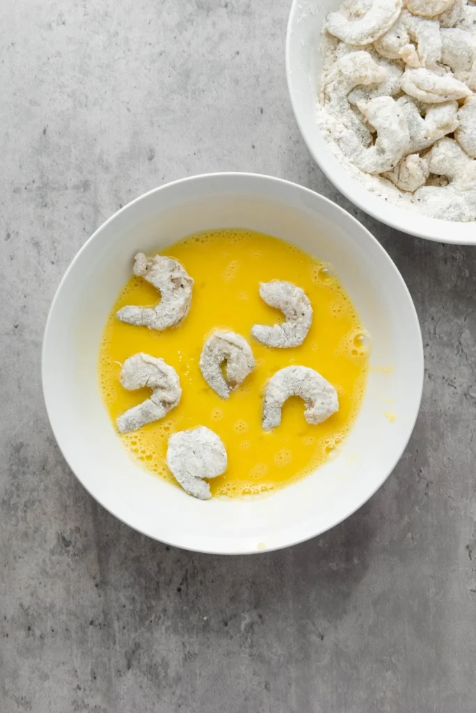 Floured shrimp being dipped into a bowl of beaten eggs.