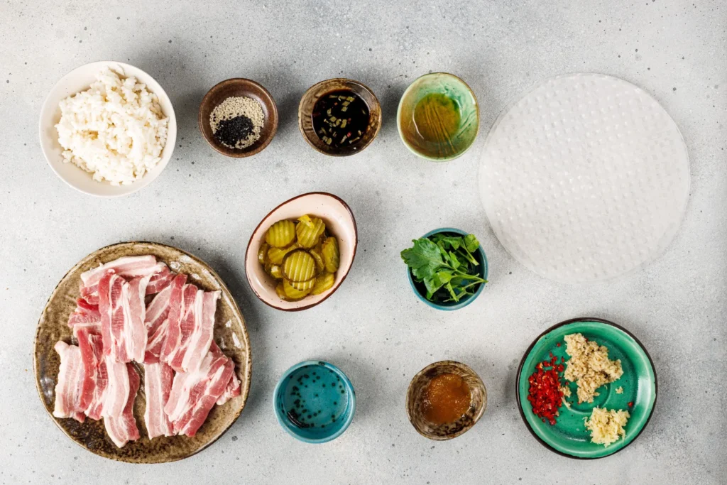 Overhead view of all ingredients for spicy pork belly hand rolls including rice, pork belly, pickled cucumbers, herbs, and rice paper.