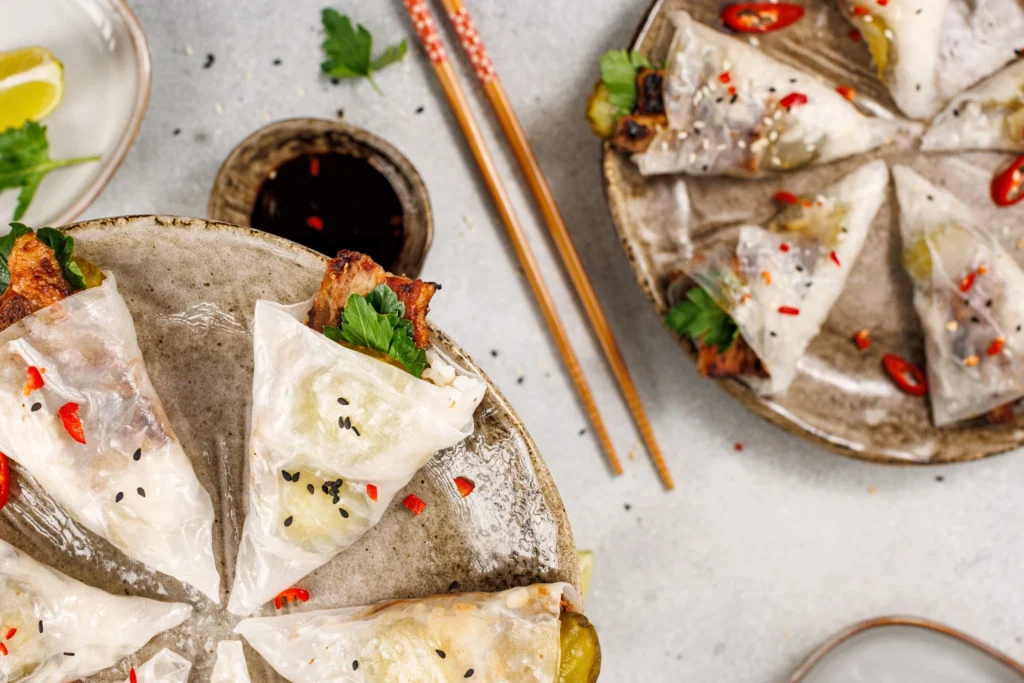 Serving platter of pork belly hand rolls in rice paper wrappers, garnished with lime wedges, sesame seeds, and chili flakes.