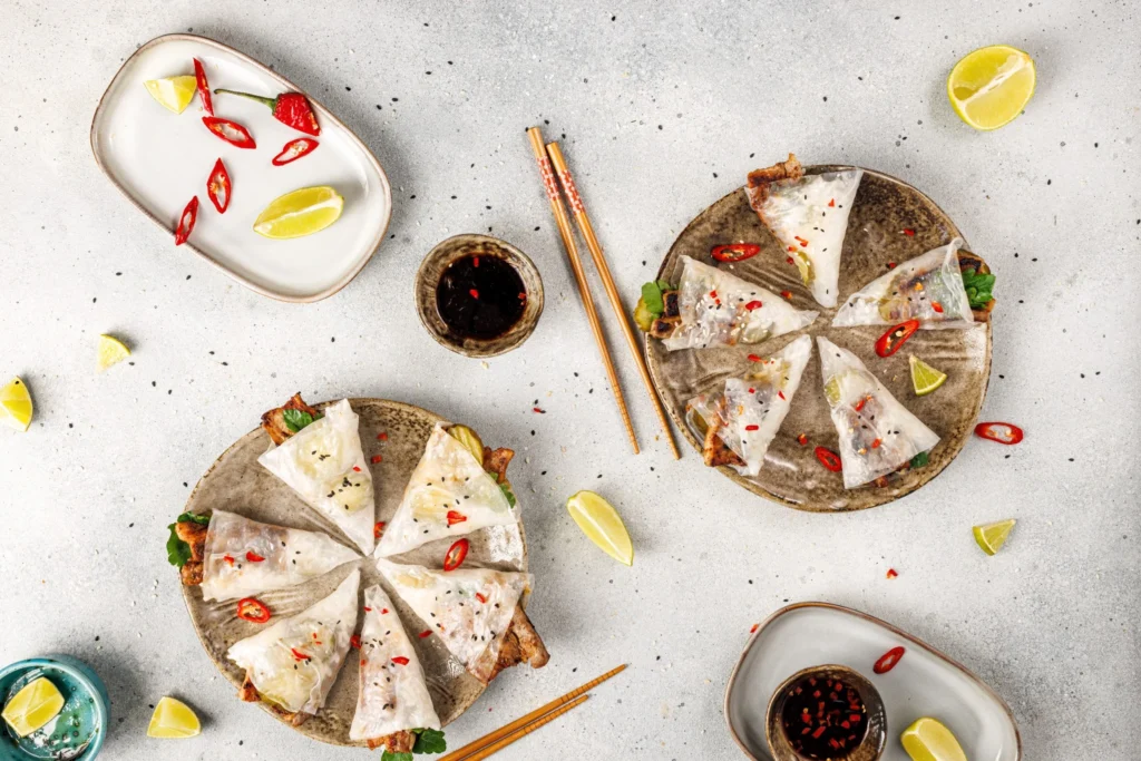 Styled flatlay of spicy pork belly hand rolls with dipping sauce, lime wedges, and chopsticks on a gray background.