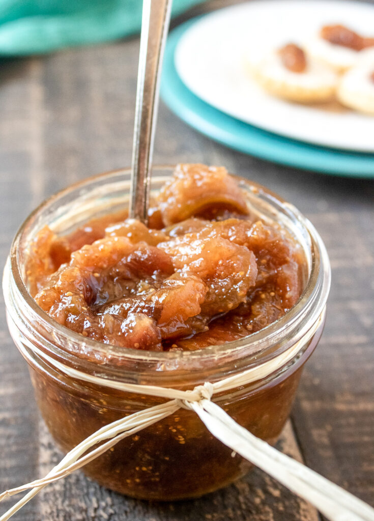 Close-up of a glass jar of vanilla fig jam with a spoon, with crackers and jam blurred in the background.