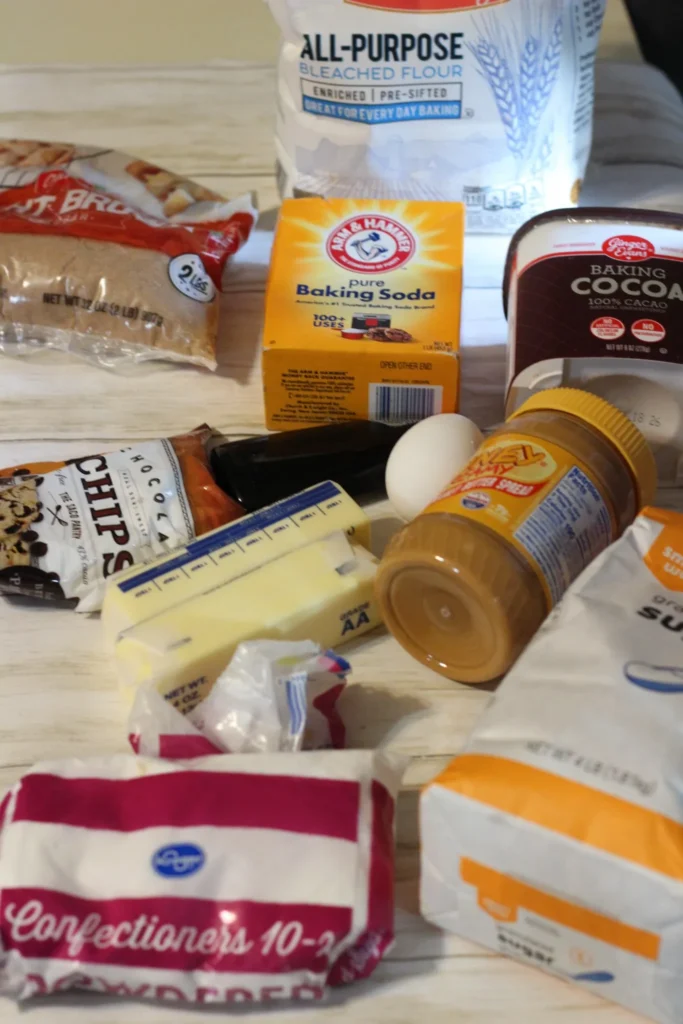 Overhead view of the ingredients for Buckeye Cookie Bars laid out on a light surface, including flour, sugars, butter, cocoa powder, peanut butter, chocolate chips, and an egg.