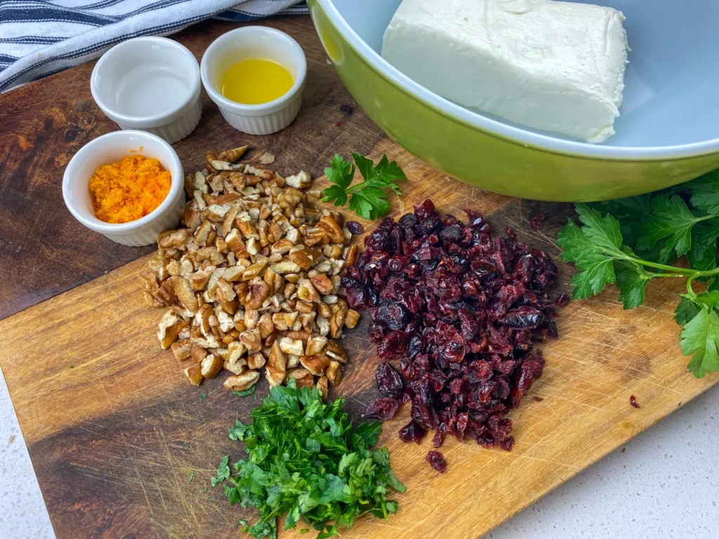 Ingredients for orange cranberry cheese ball including cream cheese, chopped pecans, dried cranberries, orange zest, parsley, and small bowls of juice and water on a wooden board.
