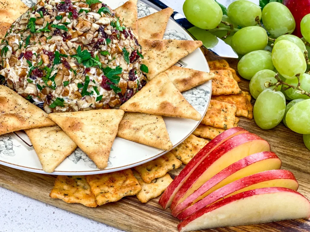 Orange cranberry cheese ball with pecan coating surrounded by pita triangles, grapes, and apple slices.
