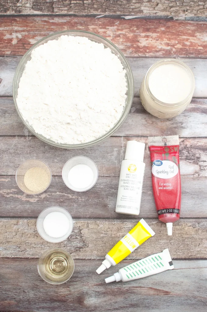 Ingredients laid out on a wooden surface, including flour, sourdough starter, yeast, sugar, salt, oil, and gel food coloring.