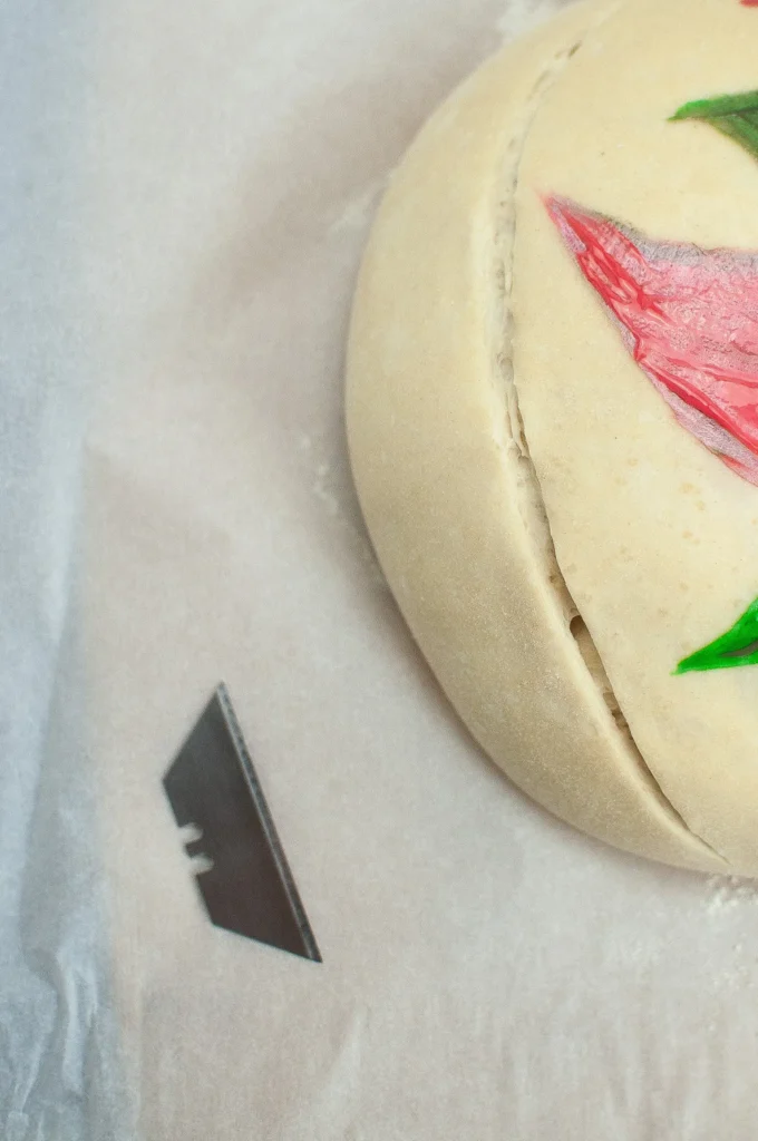 Close-up of a razor blade scoring the dough beside the hand-painted poinsettia design.