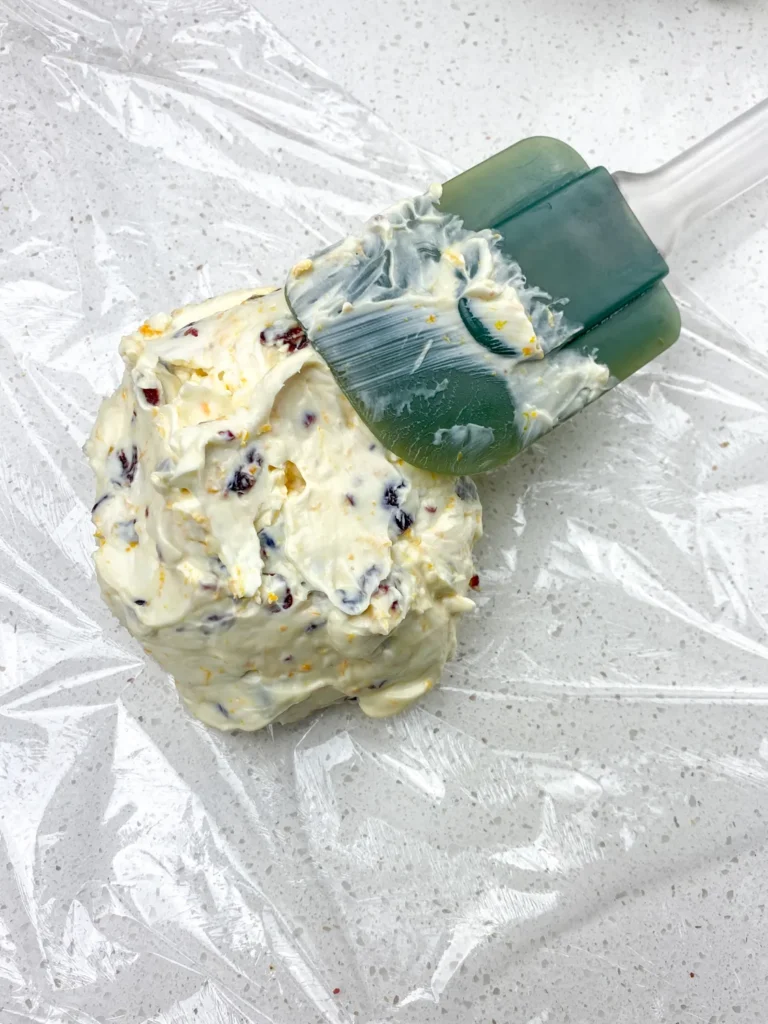 Transferring the orange cranberry cheese ball mixture onto plastic wrap using a spatula.