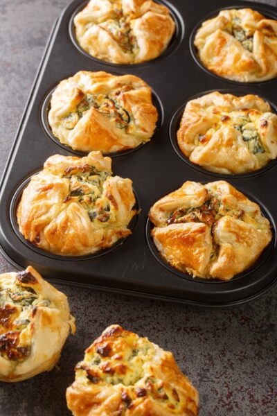 A close-up of a muffin tin with golden-brown puff pastry. The pastries are filled with spinach and cheese.