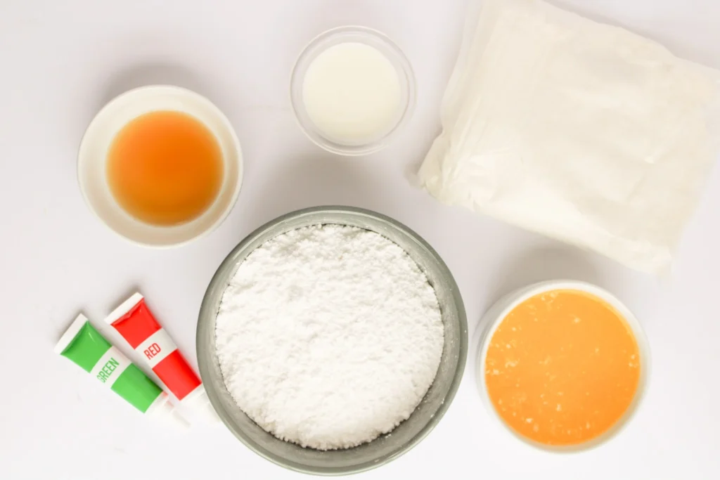 Ingredients for Christmas ornament cupcakes including powdered sugar, melted butter, vanilla, milk, and red and green food coloring on a white surface
