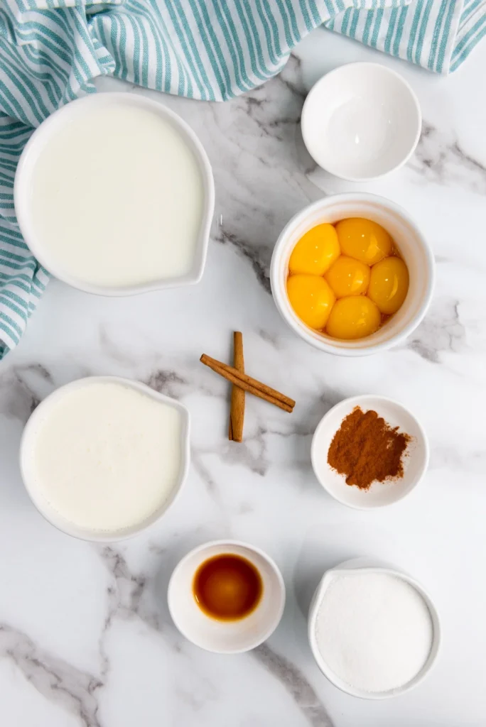 Ingredients for homemade eggnog recipe including milk, cream, egg yolks, sugar, cinnamon, and vanilla on a marble surface