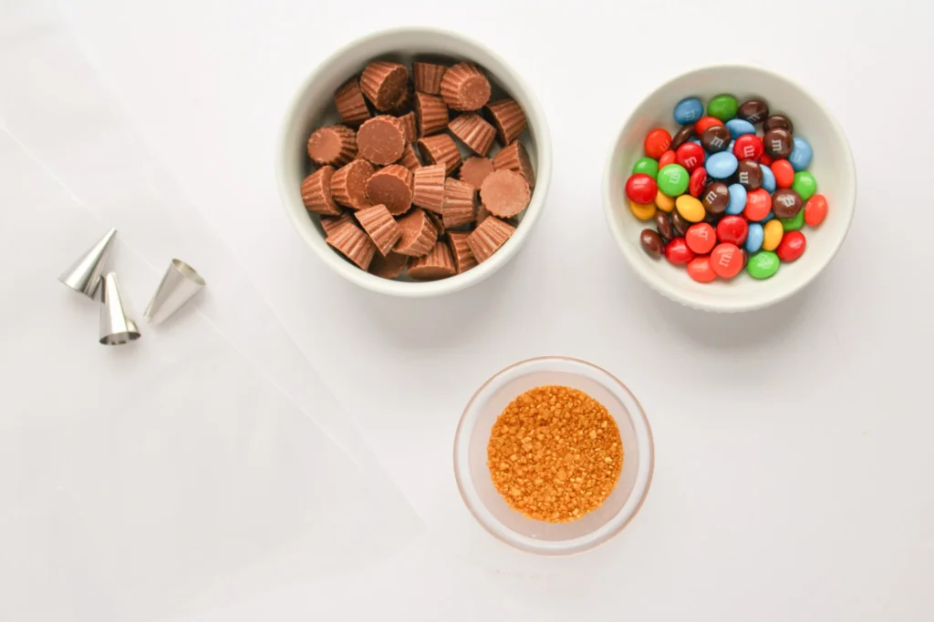 Bowls of mini Reese’s Peanut Butter Cups, red and green M&Ms, gold sanding sugar, and piping tips for decorating Christmas ornament cupcakes