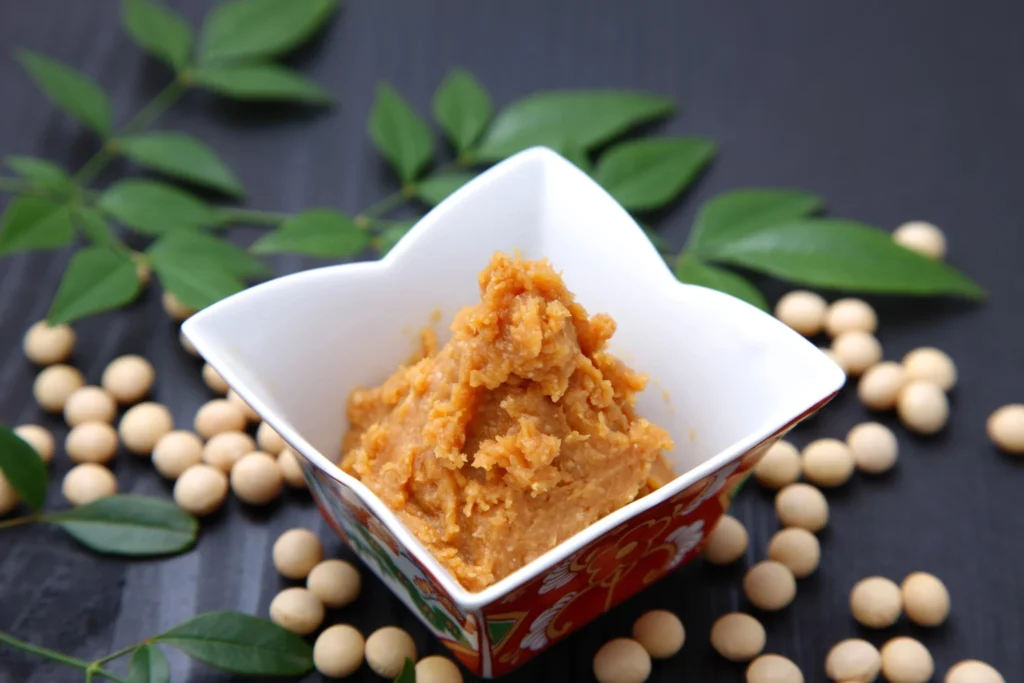 White miso paste in a decorative bowl surrounded by soybeans and green leaves