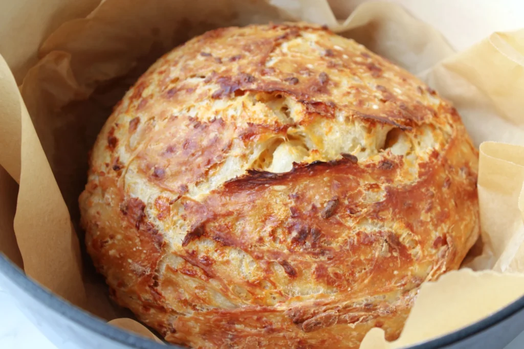 Golden brown baked no-knead cheddar bread loaf in parchment lined Dutch oven