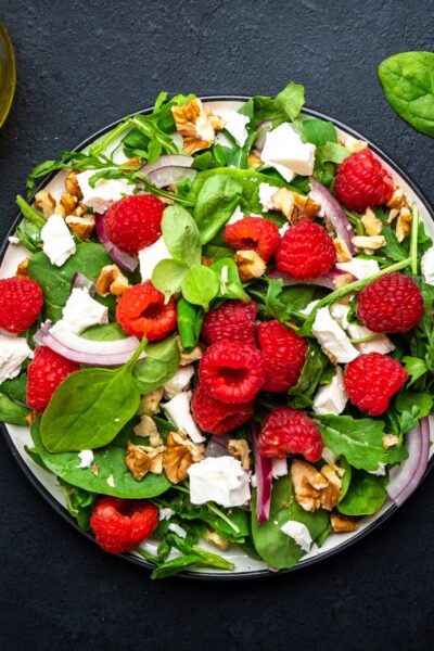 Top-view of a gourmet spring salad with raspberries, goat cheese, spinach, red onion, and nuts on a black table.