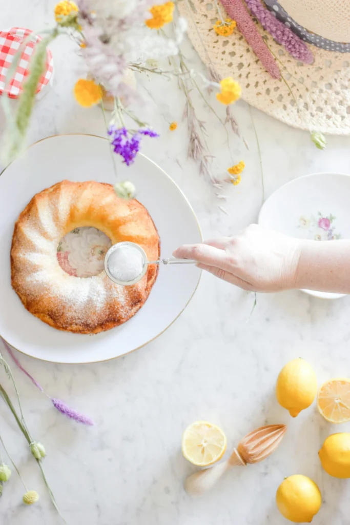Hand dusting powdered sugar over a lemon bundt cake on a plate