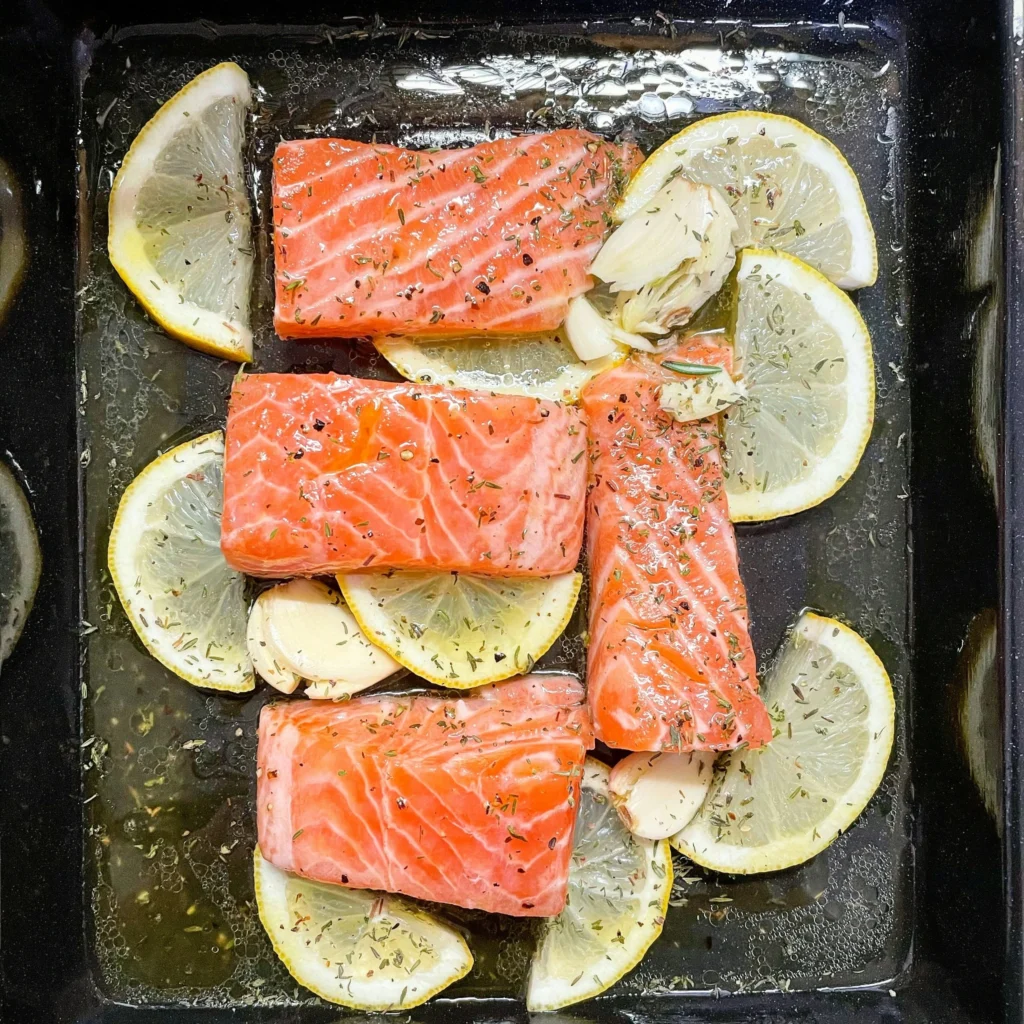 Salmon fillets baking in dish with lemon slices, garlic cloves, and herbs