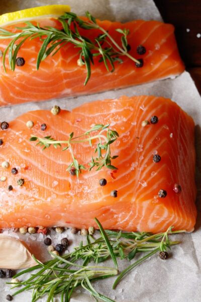 Raw salmon fillets on parchment with lemon wedges, garlic, peppercorns, and fresh herbs on a wooden surface.