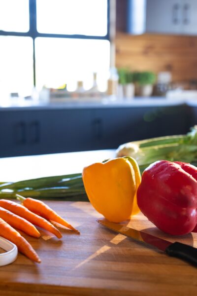 Fresh carrots, red and yellow bell peppers, and green onions on a cutting board with a knife in a bright kitchen setting.