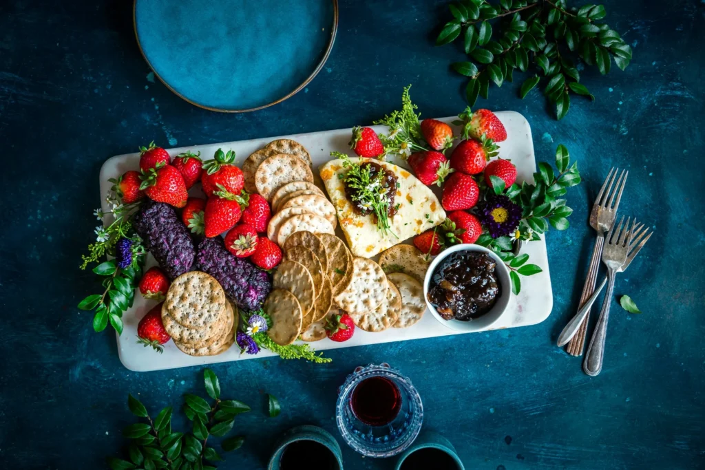 Charcuterie-style snack board with strawberries, crackers, spreads, and fresh flowers on a dark table.