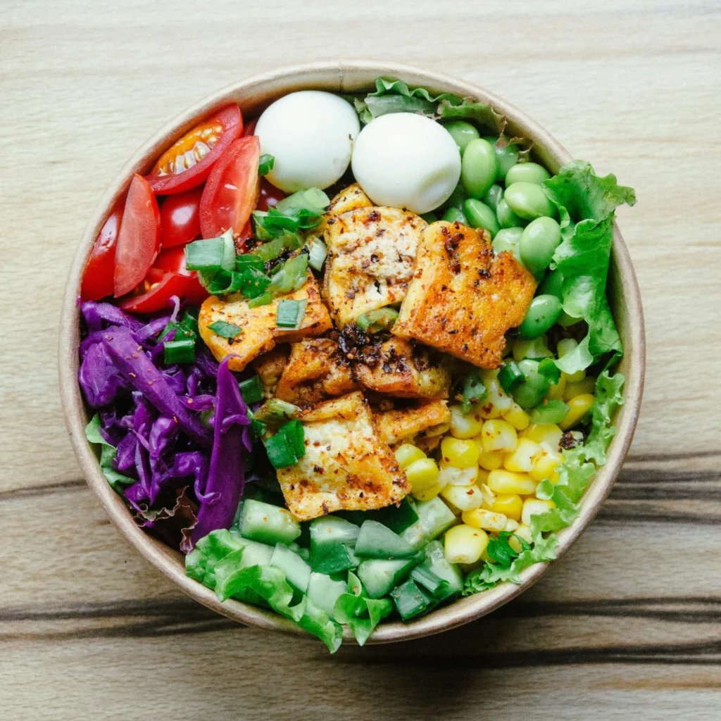 Healthy salad bowl topped with seasoned tofu, boiled eggs, tomatoes, cabbage, cucumber, corn, and edamame.