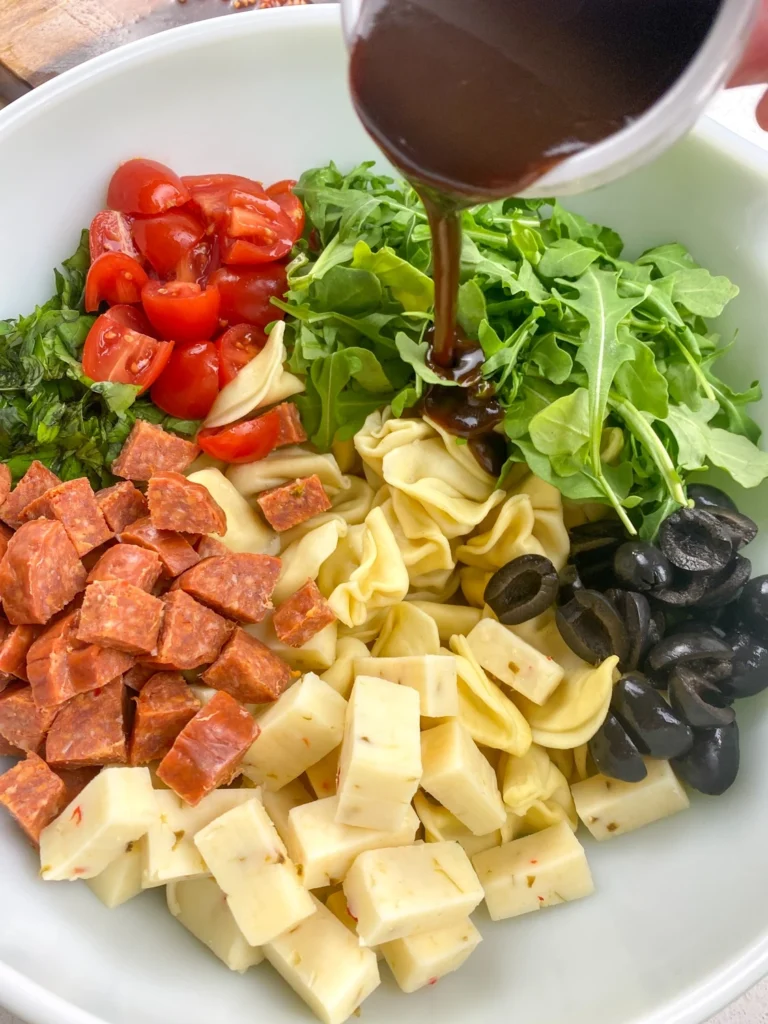 Pouring balsamic vinaigrette over tortellini antipasto salad ingredients.