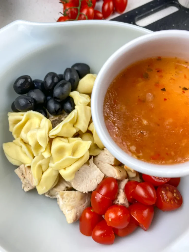 Ingredients for tortellini chicken skewers including cooked chicken, cheese tortellini, black olives, grape tomatoes, Italian dressing, and wooden skewers.