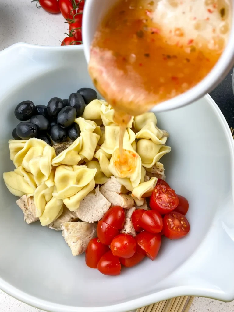 Pouring Italian dressing over tortellini, chicken, olives, and tomatoes before assembling skewers.