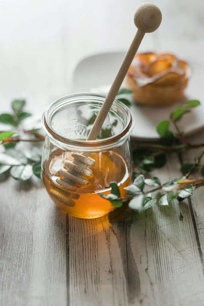 Glass jar of honey with a wooden honey dipper and pastries in the background